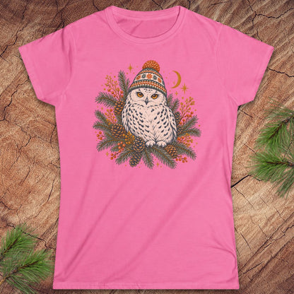 Azalea pink women's t-shirt with a Snowy Owl in an orange pom-pom knit beanie, nestled in pine boughs, pinecones, and red berries under a crescent moon, laid flat on a rustic wood background with pine branches.