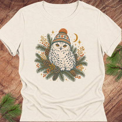 Natural cream women's relaxed t-shirt with a Snowy Owl in an orange pom-pom knit beanie, nestled in pine boughs, pinecones, and red berries under a crescent moon, laid flat on a rustic wood background with pine branches.