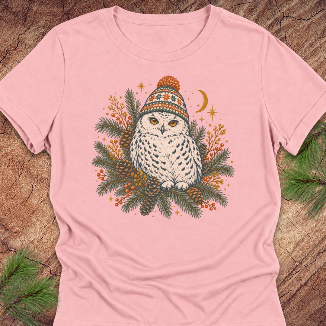 Pink women's relaxed t-shirt with a Snowy Owl in an orange pom-pom knit beanie, nestled in pine boughs, pinecones, and red berries under a crescent moon, laid flat on a rustic wood background with pine branches.