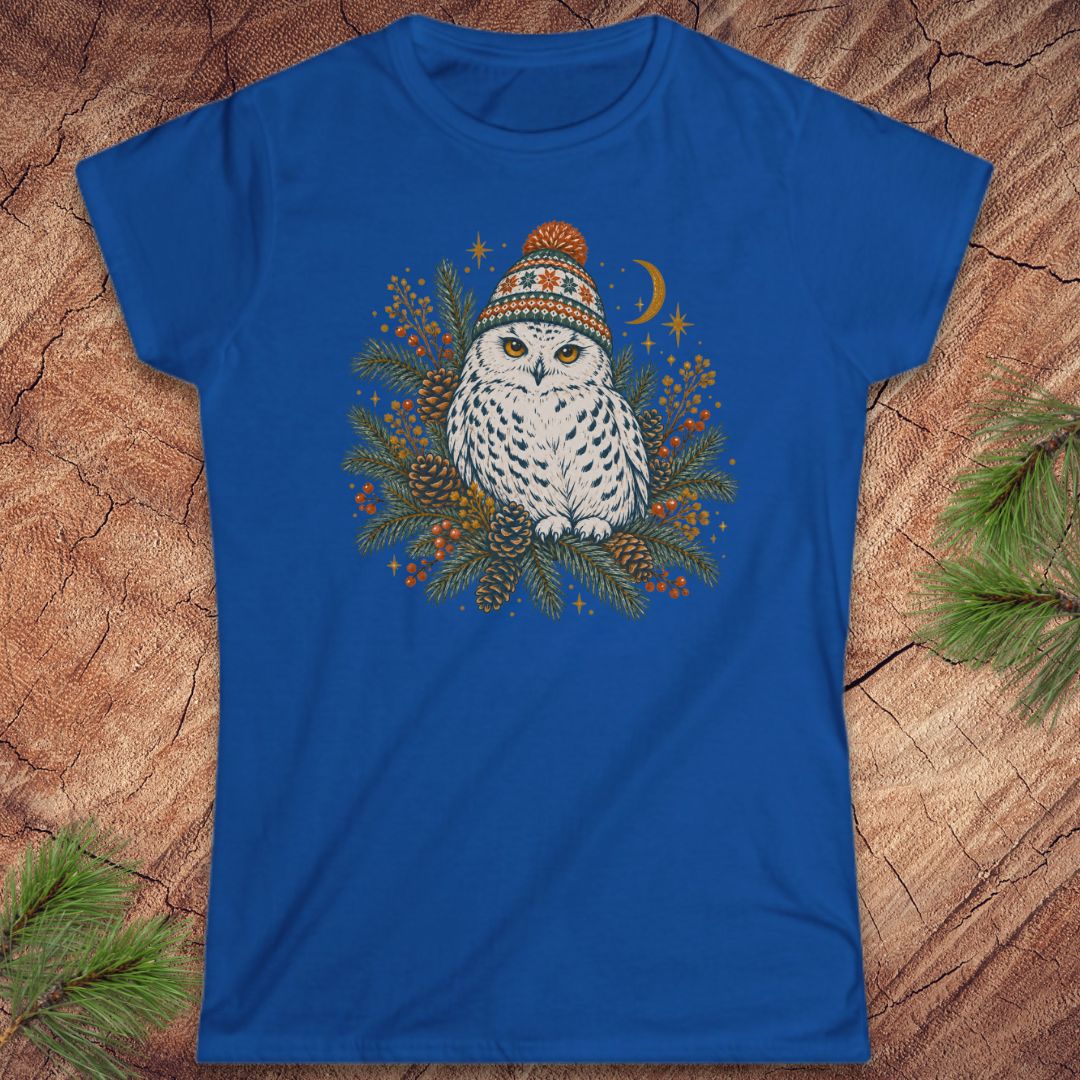 Royal blue women's t-shirt with a Snowy Owl in an orange pom-pom knit beanie, nestled in pine boughs, pinecones, and red berries under a crescent moon, laid flat on a rustic wood background with pine branches.