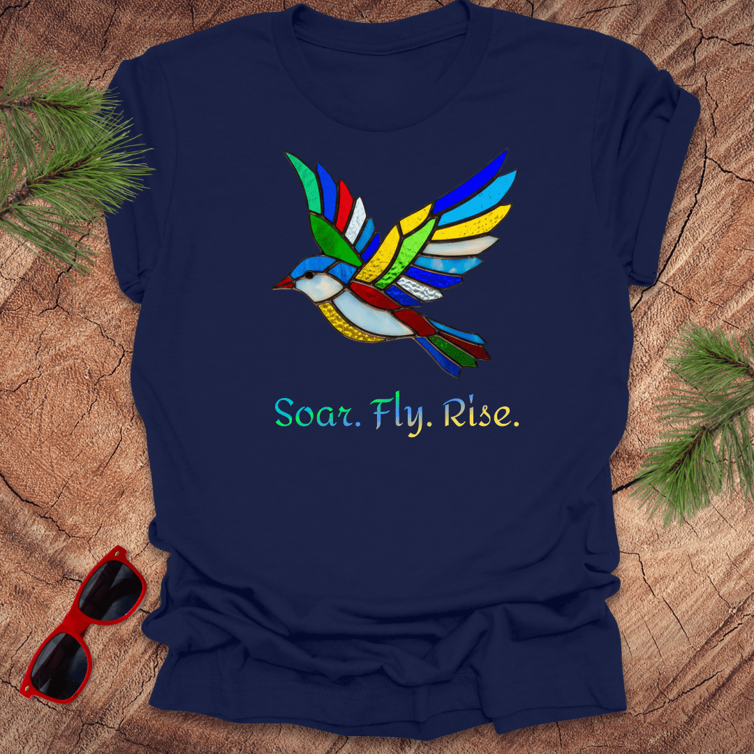 navy t-shirt with a colorful bird design and 'Soar. Fly. Rise.' text on a wooden surface with sunglasses and pine branches.