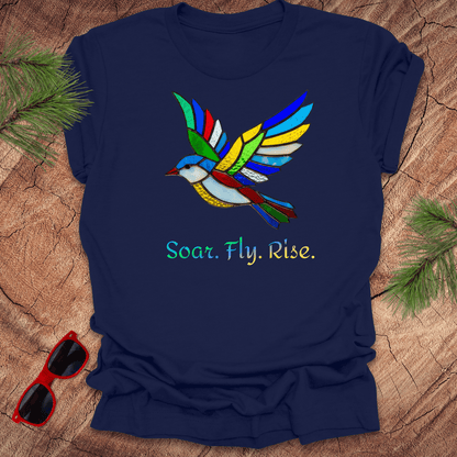 navy t-shirt with a colorful bird design and 'Soar. Fly. Rise.' text on a wooden surface with sunglasses and pine branches.