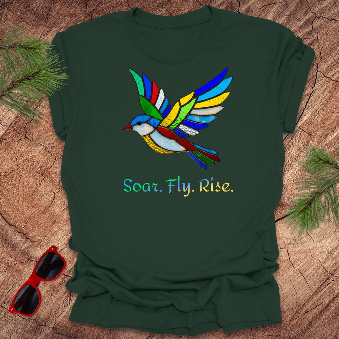 Green t-shirt with a colorful bird design and 'Soar. Fly. Rise.' text on a wooden surface with sunglasses and pine branches.