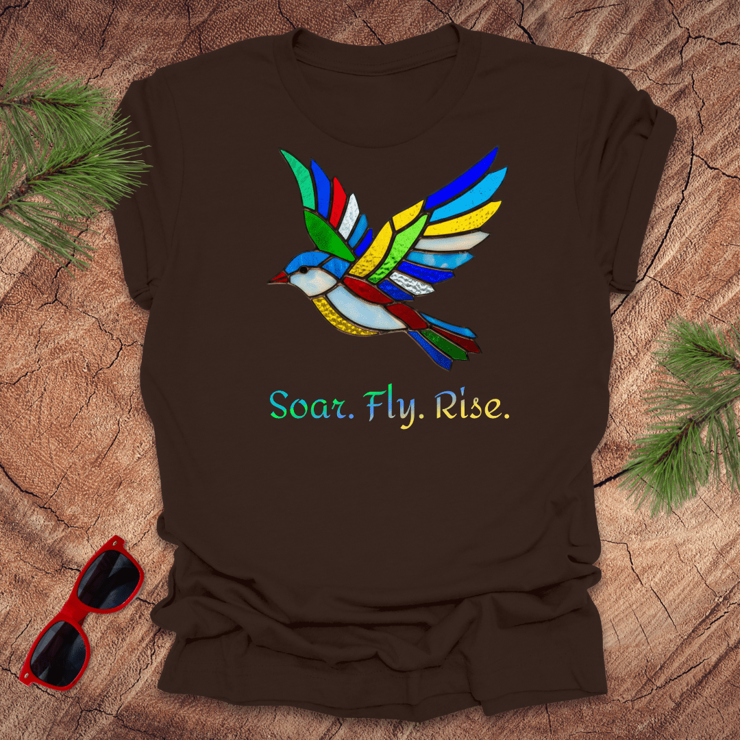 Brown t-shirt with colorful bird design and 'Soar. Fly. Rise.' text on a wooden surface with sunglasses and pine branches.