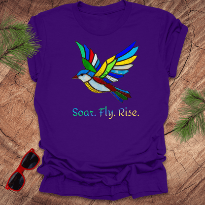 Purple t-shirt with a colorful bird design and 'Soar. Fly. Rise.' text on a wooden surface.