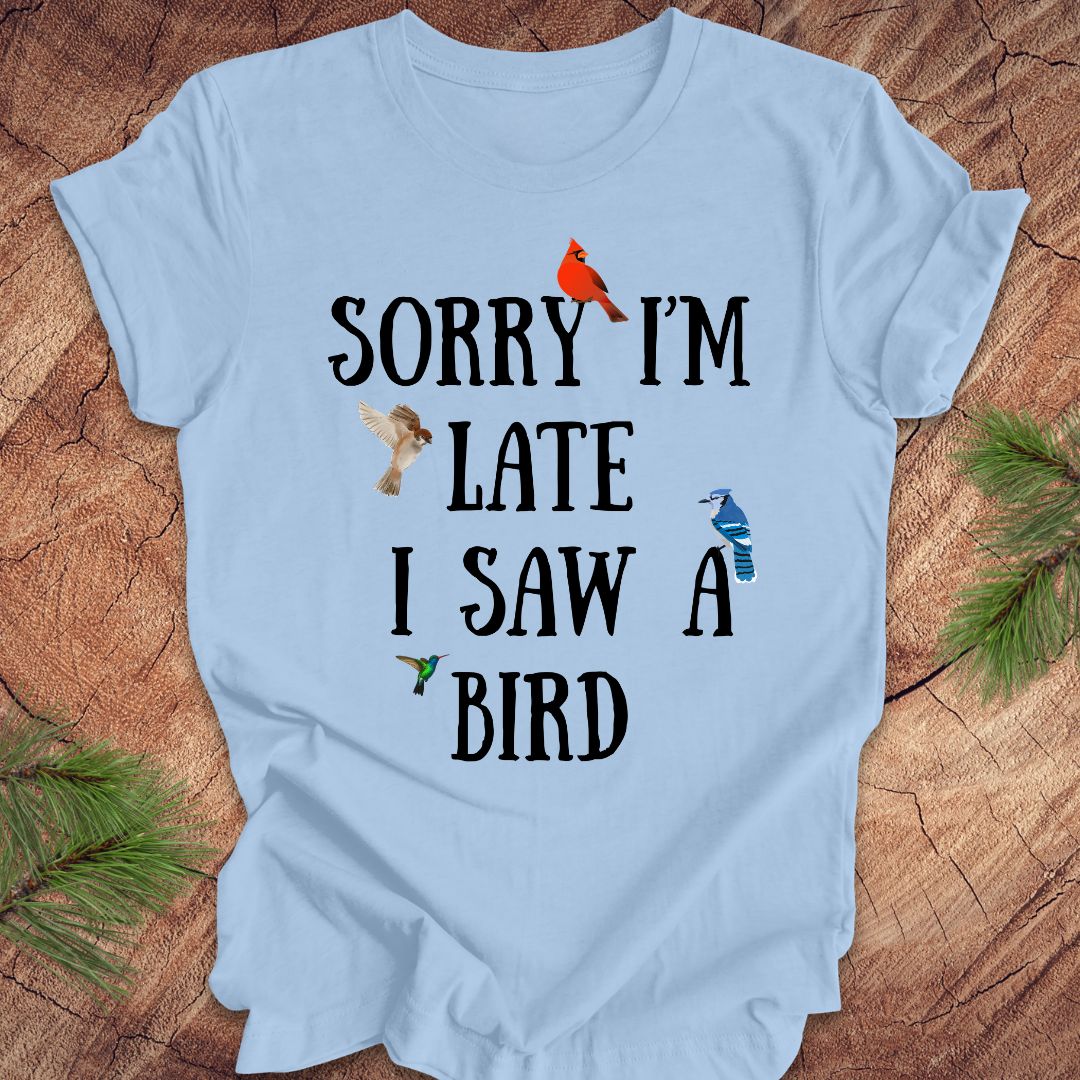 Light blue t-shirt with text 'Sorry I'm late I saw a bird' on a wooden surface.