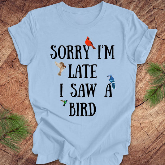 Light blue t-shirt with text 'Sorry I'm late I saw a bird' on a wooden surface.
