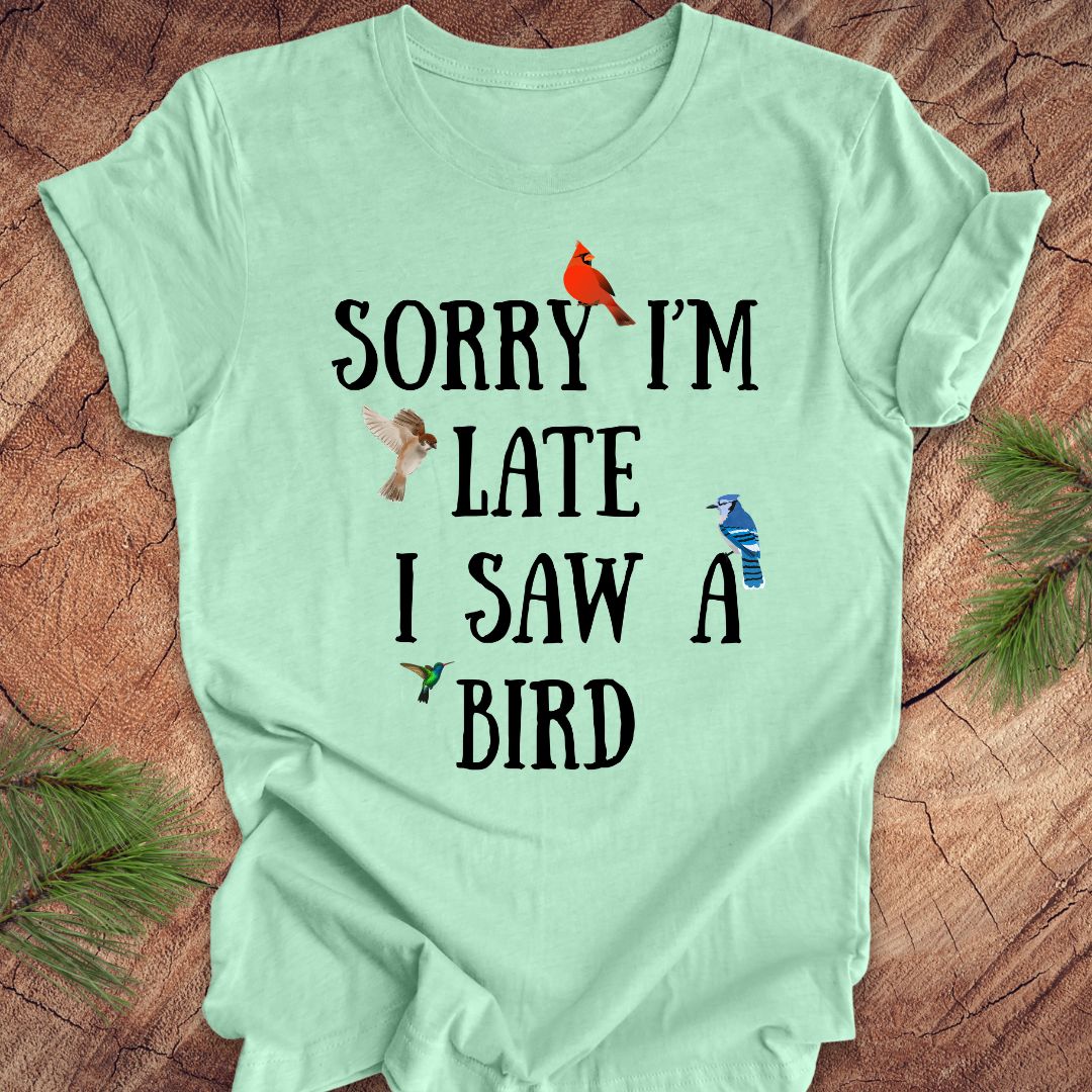 Light green t-shirt with text 'Sorry I'm late I saw a bird' on a wooden surface with decorative elements.