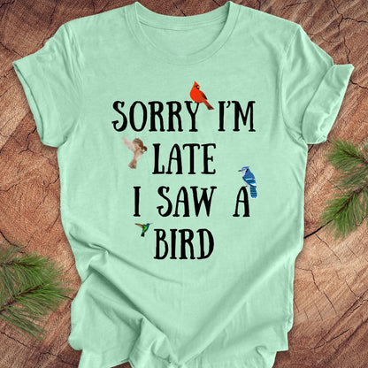 Light green t-shirt with text 'Sorry I'm late I saw a bird' on a wooden surface with decorative elements.