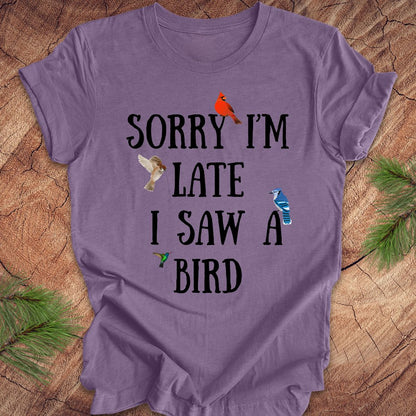 Purple t-shirt with text 'Sorry I'm late I saw a bird' on a wooden surface.