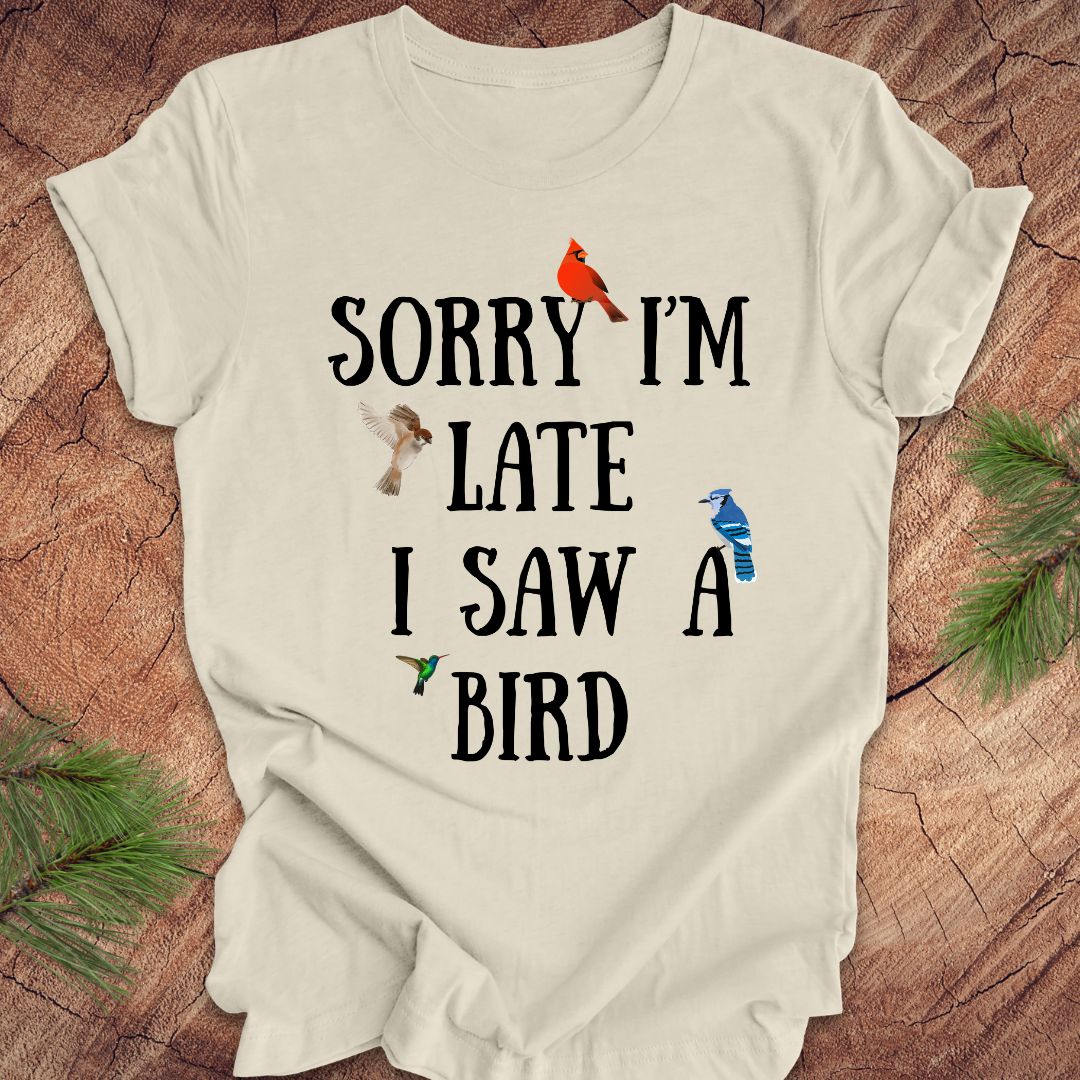 Beige t-shirt with text 'Sorry I'm late I saw a bird' on a wooden surface with decorative elements.