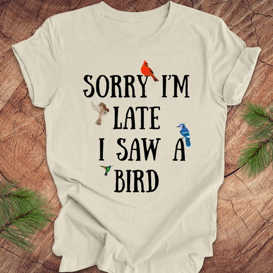 Beige t-shirt with text 'Sorry I'm late I saw a bird' on a wooden surface with decorative elements.