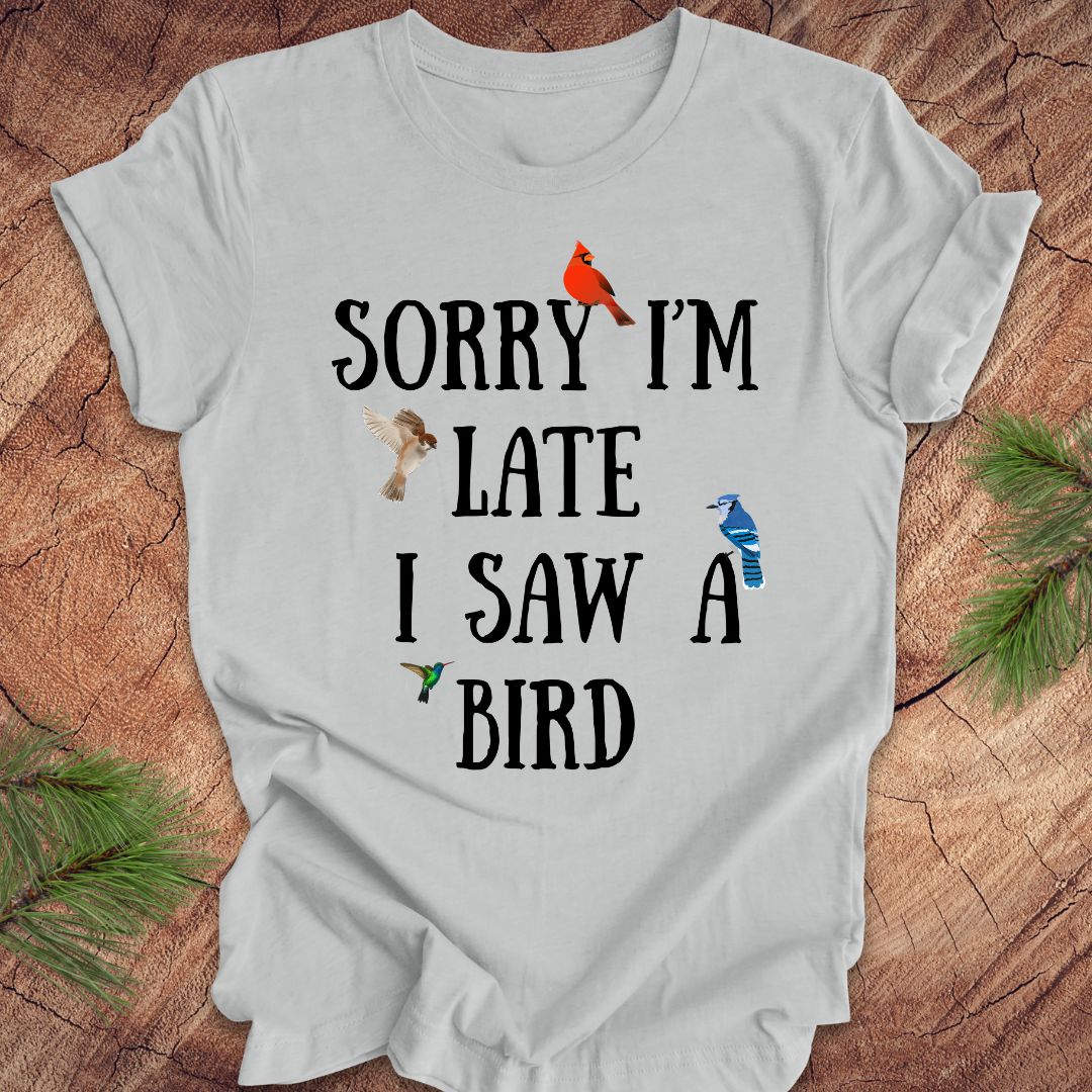 Gray t-shirt with text 'Sorry I'm late I saw a bird' on a wooden surface with decorative elements.