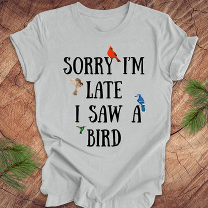 Gray t-shirt with text 'Sorry I'm late I saw a bird' on a wooden surface with decorative elements.