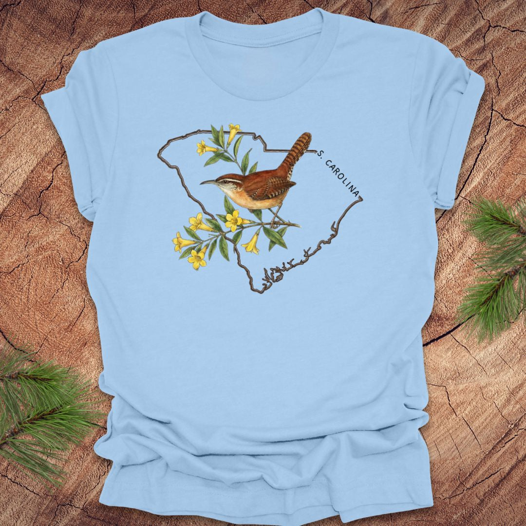 baby blue t-shirt with a carolina wren and yellow jessamine design on an outline of south carolina, placed on a wooden surface.