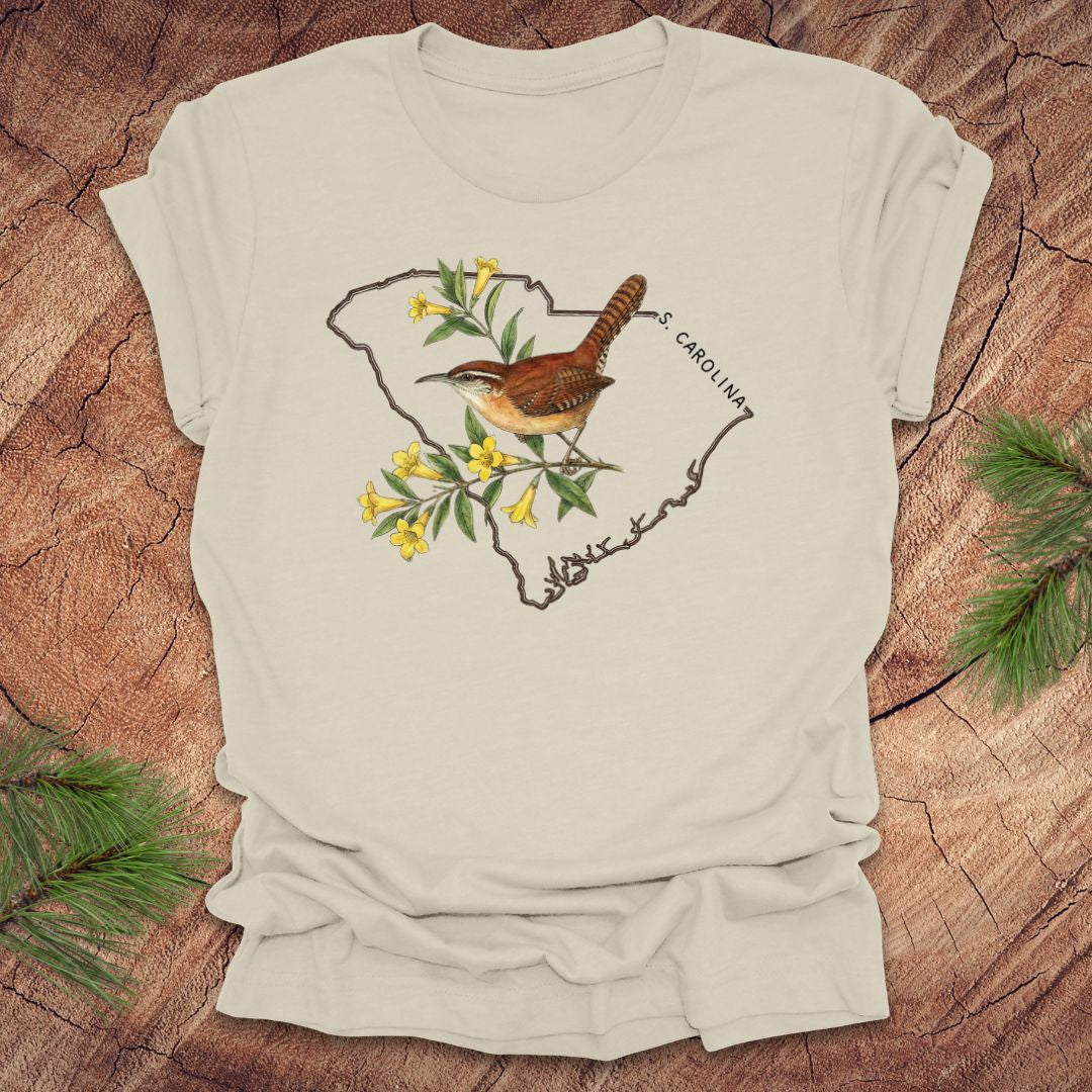 Beige t-shirt with a carolina wren and yellow jessamine design on an outline of south carolina, placed on a wooden surface.