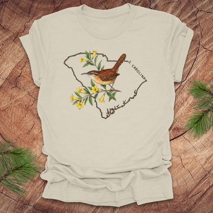 Beige t-shirt with a carolina wren and yellow jessamine design on an outline of south carolina, placed on a wooden surface.