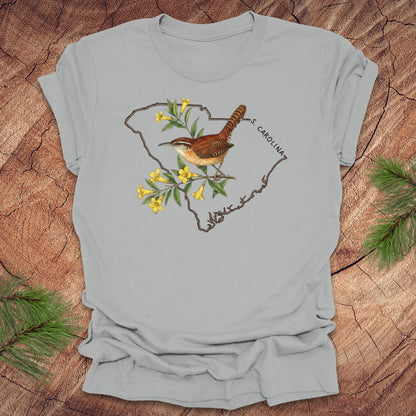 Silver t-shirt with a carolina wren and yellow jessamine design on an outline of south carolina, placed on a wooden surface.