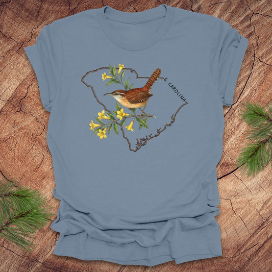 Steel blue t-shirt with a carolina wren and yellow jessamine design on an outline of south carolina, placed on a wooden surface.
