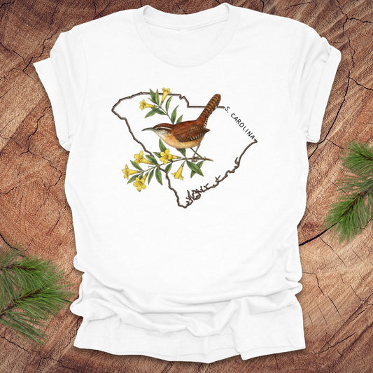 White t-shirt with a carolina wren and yellow jessamine design on an outline of south carolina, placed on a wooden surface.