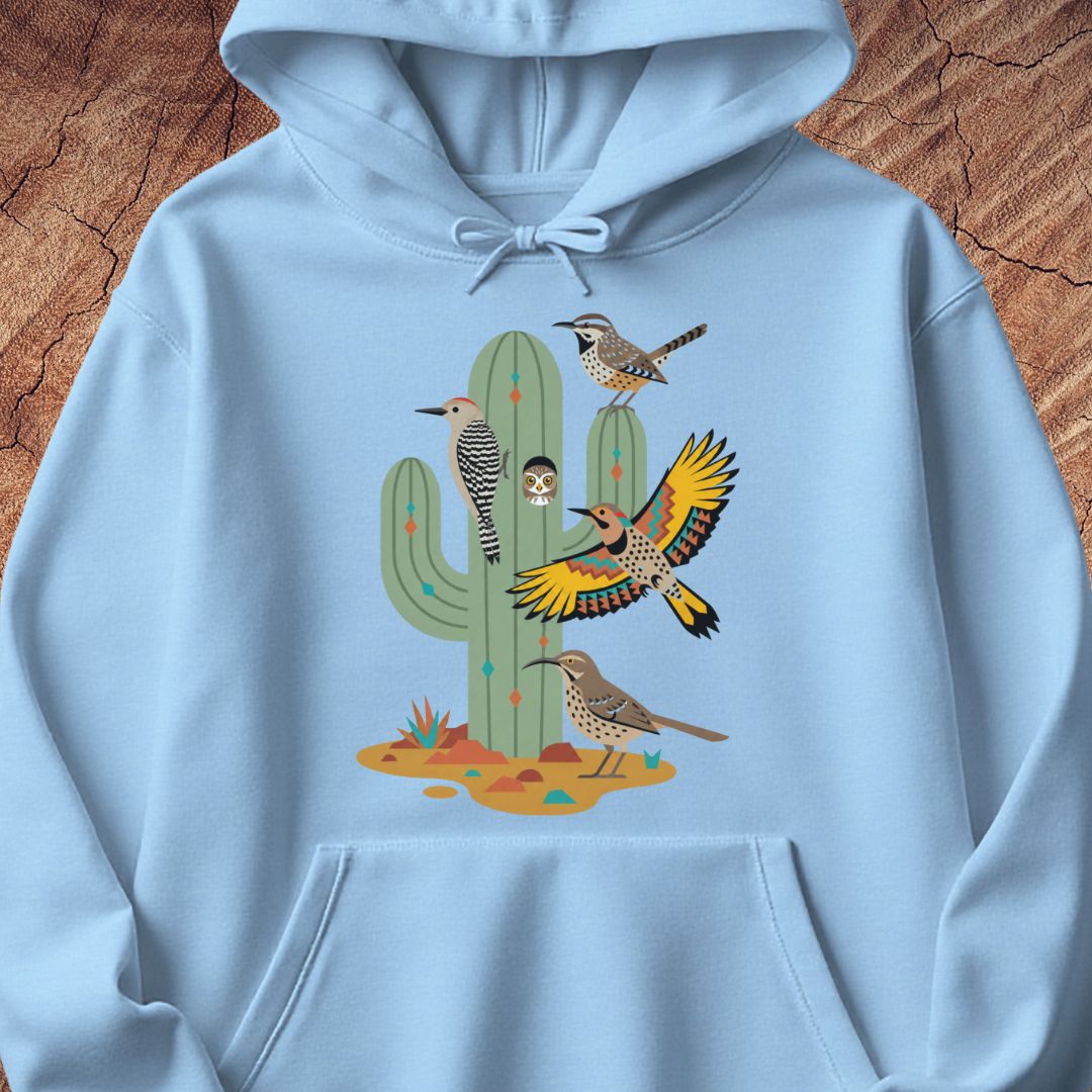 A light blue hoodie laid flat on a textured brown wood background, featuring a colorful graphic print of desert birds and a saguaro cactus on the front.