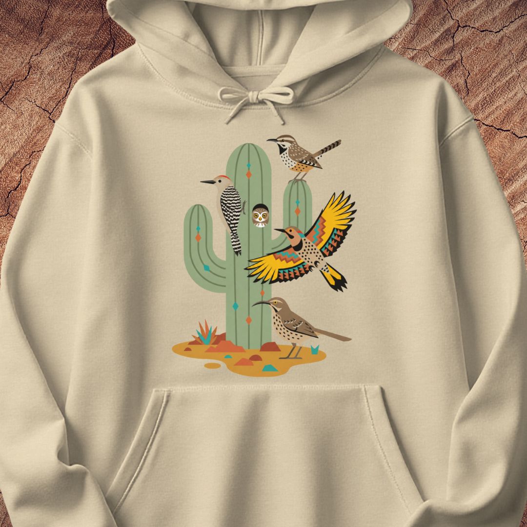 A cream-colored pullover hoodie featuring a colorful graphic print of desert birds and a saguaro cactus, displayed on a textured brown wood background.