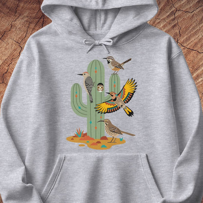 A light gray pullover hoodie featuring a colorful graphic print of desert birds perched on and around a saguaro cactus, displayed flat against a rustic wood background.
