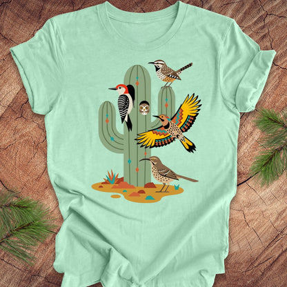Mint green t-shirt with a cactus and birds design on a wooden surface.