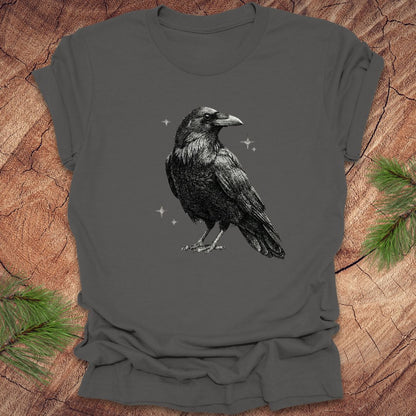 Gray t-shirt with a black raven design on a wooden surface with pine branches.