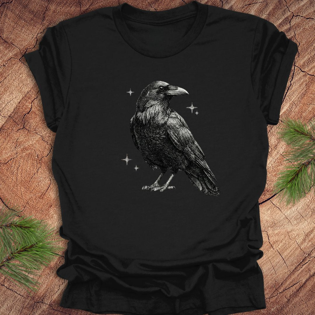 Black t-shirt with a raven design on a wooden surface with pine branches.