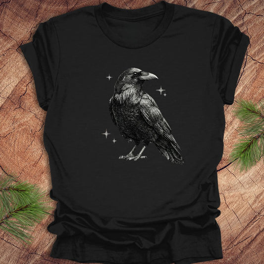 Black t-shirt with a raven design on a wooden surface with pine branches.