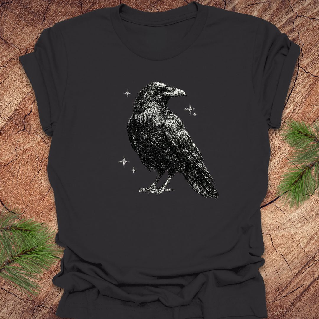 Dark gray t-shirt with a raven design on a wooden surface with pine branches.