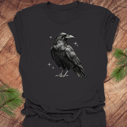 Dark gray t-shirt with a raven design on a wooden surface with pine branches.