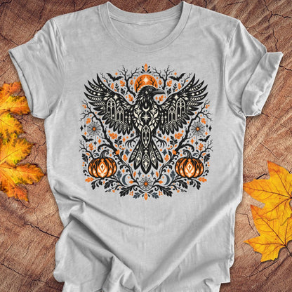 A gray heather t-shirt with a folk art crow and pumpkin halloween design on a wood background with fall leaves. 