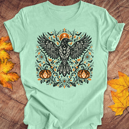 A mint green t-shirt with a folk art crow and pumpkin halloween design on a wood background with fall leaves. 