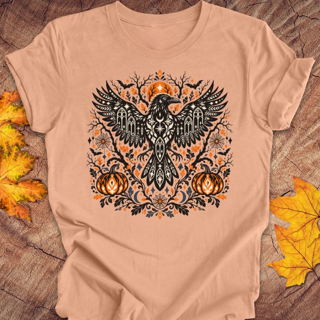 A peach t-shirt with a folk art crow and pumpkin halloween design on a wood background with fall leaves. 