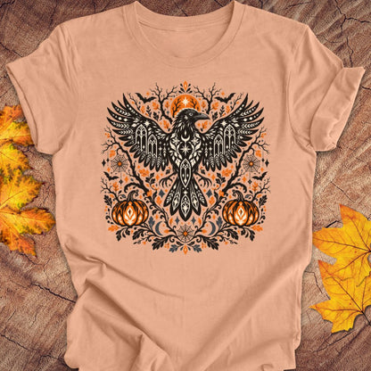 A peach t-shirt with a folk art crow and pumpkin halloween design on a wood background with fall leaves. 