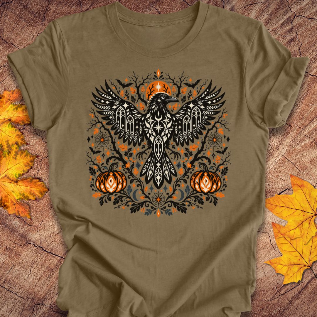 An olive green t-shirt with a folk art crow and pumpkin halloween design on a wood background with fall leaves. 