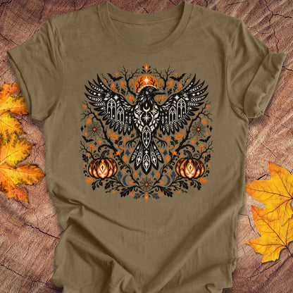 An olive green t-shirt with a folk art crow and pumpkin halloween design on a wood background with fall leaves. 