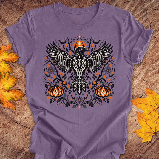 A purple t-shirt with a folk art crow and pumpkin halloween design on a wood background with fall leaves. 