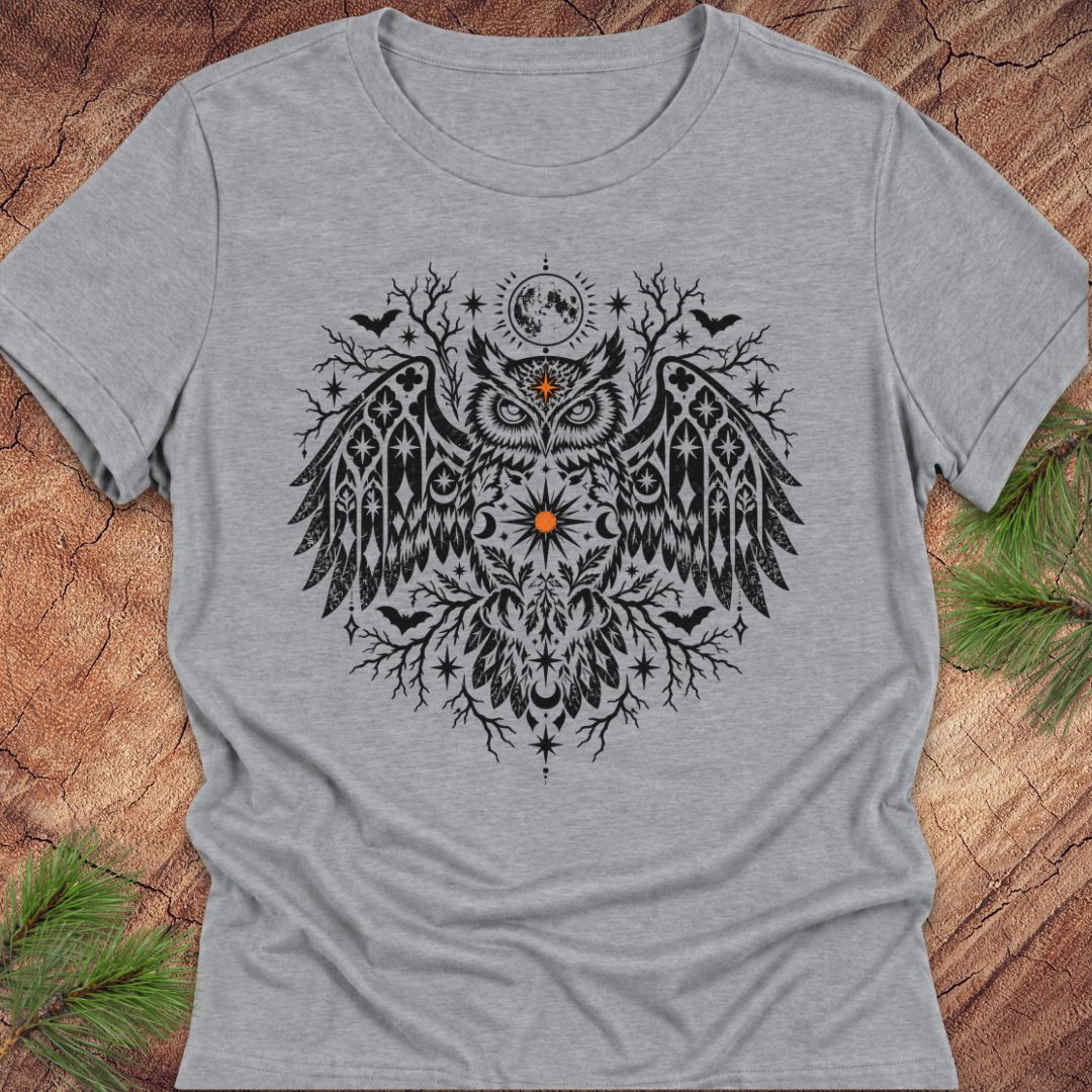 A heather gray short-sleeve t-shirt with a detailed black owl graphic on the front, featuring a moon and stars motif, laid flat on a rustic wood background with pine needles in the corners.