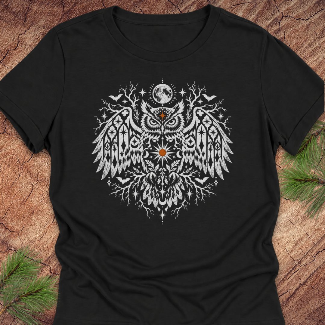 A black short-sleeve crewneck t-shirt featuring a large white owl graphic with intricate tribal-style patterns, a moon and stars above, and a small orange accent in the center. The shirt is laid flat on a rustic wood background with pine needles in the corners.
