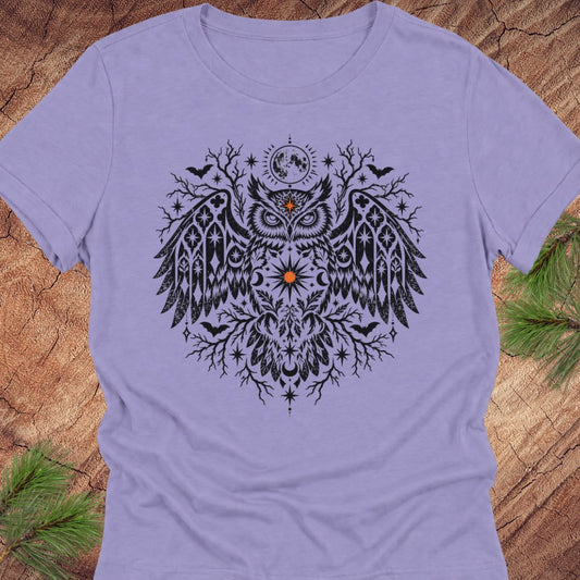A lavender short-sleeve crewneck t-shirt featuring a detailed black owl graphic with tribal patterns, a crescent moon, and stars, laid flat on a rustic wood background with pine needles.