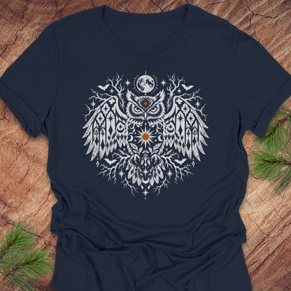 A navy blue short-sleeve t-shirt with a detailed white owl graphic on the chest, laid flat on a rustic wood background with pine branches in the corners.