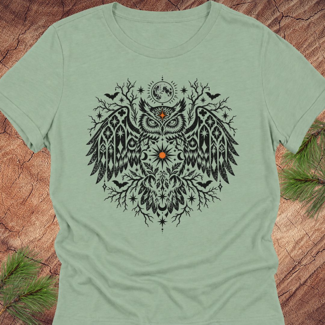 A sage green short-sleeve crewneck t-shirt featuring a large black owl graphic with intricate tribal patterns, a crescent moon, and stars on the chest. The t-shirt is laid flat on a rustic wood background with pine needles visible in the corners.