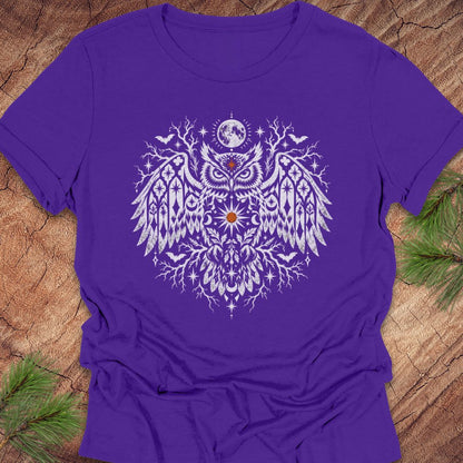 A purple short-sleeve crewneck t-shirt featuring a large white owl graphic with intricate tribal-style patterns, a crescent moon, and stars on the chest. The shirt is laid flat on a rustic wood background with pine needles visible in the corners.