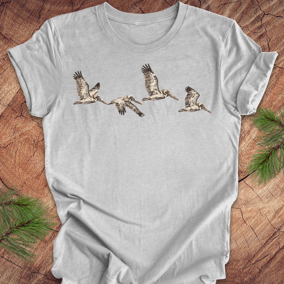 A light gray short-sleeve t-shirt with a graphic print of five brown pelicans flying in a row, laid flat on a rustic wood background with pine needles in the corners.