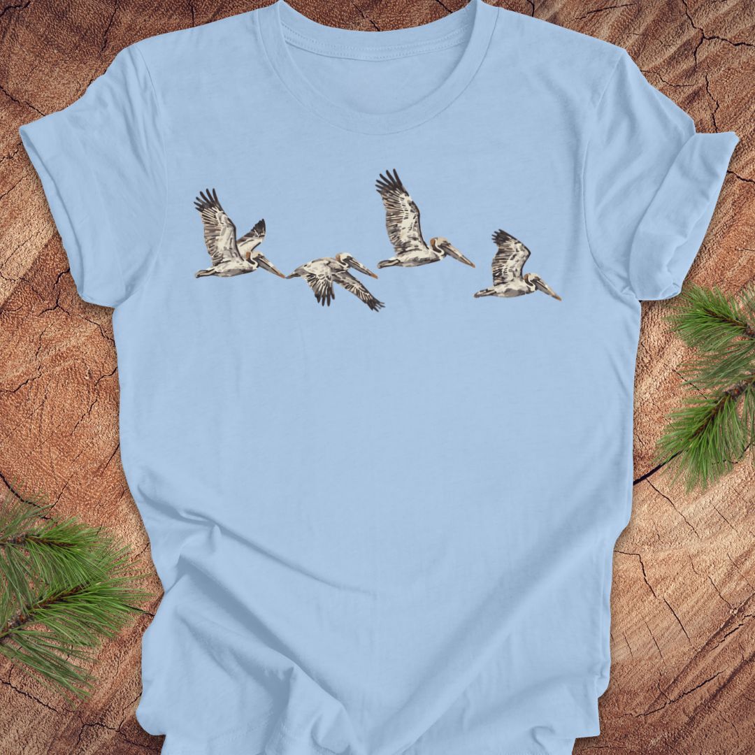 A light blue short-sleeve t-shirt laid flat on a rustic wood surface, featuring a graphic print of five flying pelicans across the chest. Green pine branches are placed in the corners as styling props.