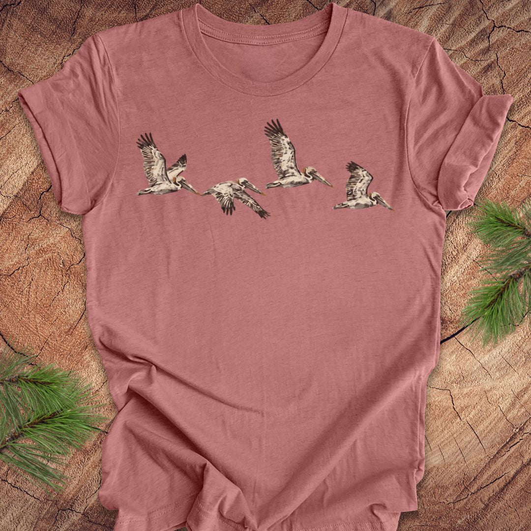A dusty rose short-sleeve t-shirt featuring a graphic print of five flying birds, laid flat on a rustic wood background with pine needles in the corners.