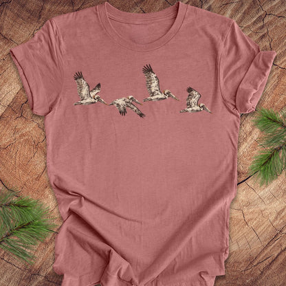 A dusty rose short-sleeve t-shirt featuring a graphic print of five flying birds, laid flat on a rustic wood background with pine needles in the corners.